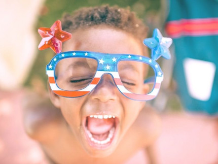 Child in red white and blue fourth of july novelty glasses looking up at the camera with a big smile